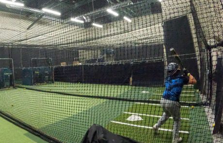 Youth baseball player swinging inside an indoor batting cage with pitching machine, practicing timing and contact on turf