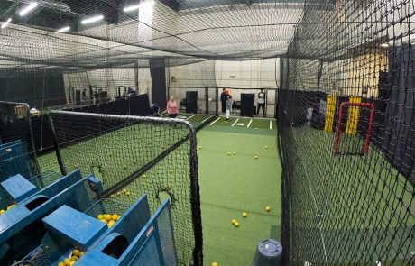 Wide view of indoor batting cages with multiple lanes, pitching machines, and players training simultaneously on turf fields